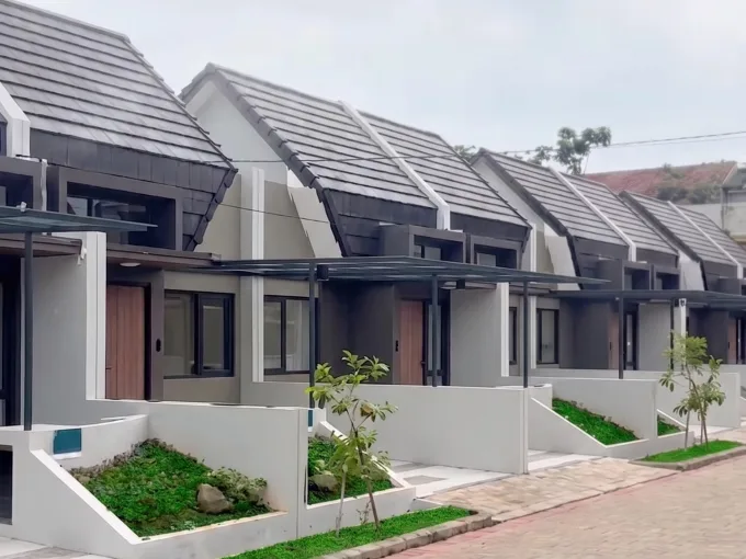 Row of modern, single-story townhouses with gray tiled roofs and small landscaped front yards along a paved street, under a cloudy sky, with a 'GARDENS at Candi Sawan' logo above them.