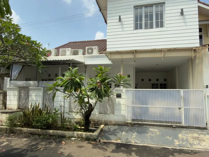 White two‑story house with a white metal gate, a small front garden with a flowering tree, and several air conditioning units on the roof against a blue sky.