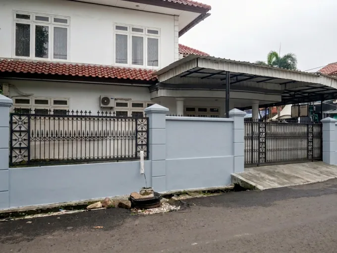 Front view of a two-story house behind a pale blue boundary wall and decorative black wrought-iron fence with a red tile roof and carport.