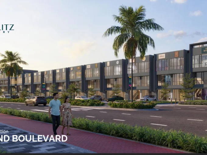 Promotional image of The Floritz Gallery Grand Boulevard shopping complex with palm trees and a couple walking on a red brick sidewalk along a multi‑storey plaza road at dusk.