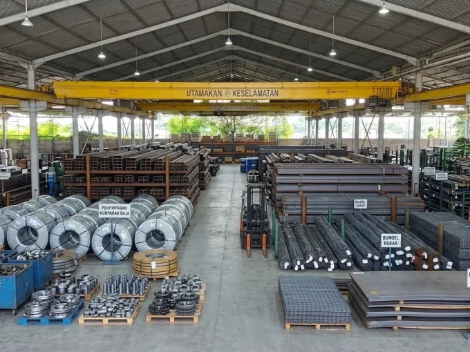 Industrial warehouse with neatly stacked steel pipes, coils, and metal sheets; a forklift sits in the central aisle.